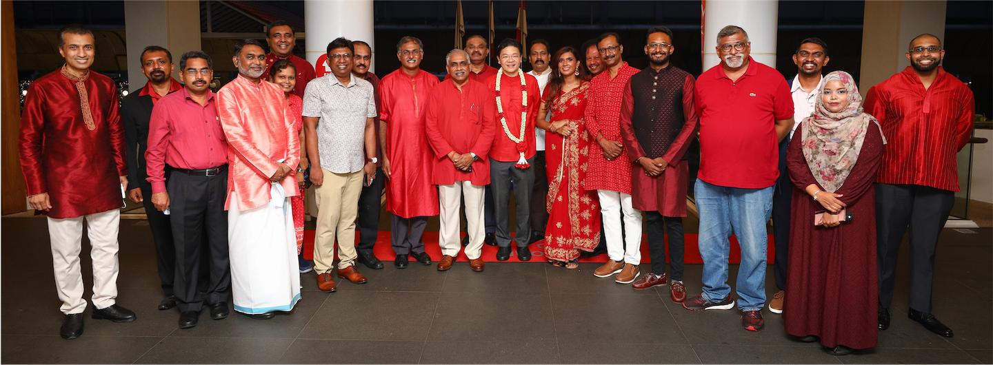 Group of people in red attire, some wearing traditional clothing and flower garlands, standing indoors.